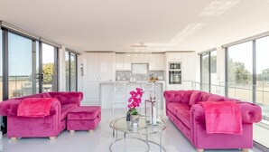 Ein modernes Wohnzimmer mit zwei rosafarbenen Sofas, einem Glastisch und einer Küche im Hintergrund.