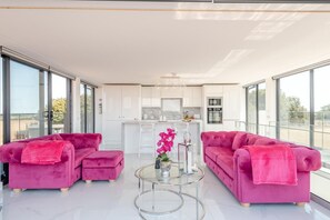 Ein modernes Wohnzimmer mit zwei rosafarbenen Sofas, einem Glastisch und einer Küche im Hintergrund.