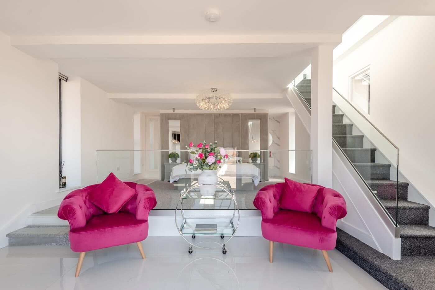 A modern living room with a glass coffee table, two pink armchairs, a chandelier, and a staircase.