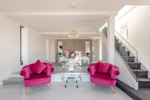 A modern living room with a glass coffee table, two pink armchairs, a chandelier, and a staircase.