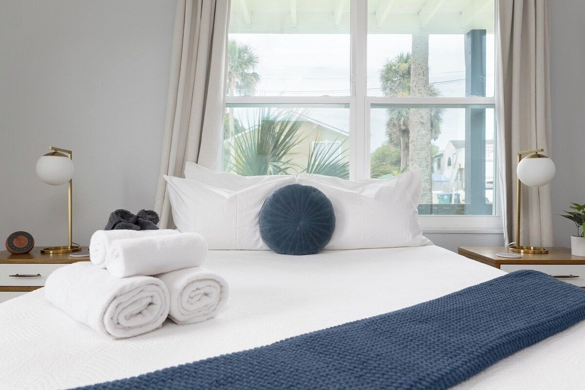 Uma cama bem arrumada com lençóis brancos, um cobertor azul e um travesseiro azul. Há duas mesas de cabeceira com luminárias e um pequeno relógio em uma delas.