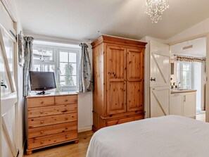 Une chambre à coucher avec une commode en bois, un lit, un téléviseur posé sur une commode et une fenêtre avec des rideaux.