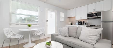 A modern, well-lit kitchen with white cabinets, a stainless steel refrigerator, and a microwave. There is a gray sofa with pillows, a white round table with a plant, and two white chairs.
