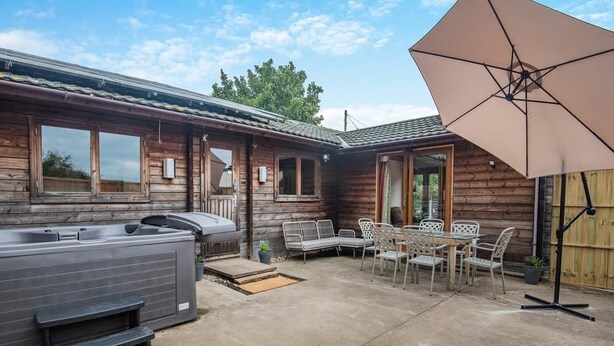 Een houten hut met een hottub, terrasmeubels en een grote parasol.