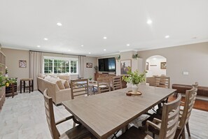 Un vaste espace dédié à la salle à manger, avec une grande table en bois et de nombreuses chaises, un ensemble de canapés et un téléviseur en arrière-plan.