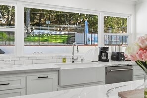 A modern kitchen with a large window, white cabinetry, and a view of a fenced outdoor area. - Remodeled 4BR Retreat - FirePit + Basketball Court (Atascadero)