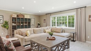 Ein Wohnzimmer mit Ecksofa, Couchtisch, Bücherregal und einem großen Fenster mit Vorhängen.