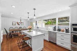 A modern kitchen with white cabinetry, a central island with bar stools, a dishwasher, and a view of the outdoors through large windows. - Remodeled 4BR Retreat - FirePit + Basketball Court (Atascadero)