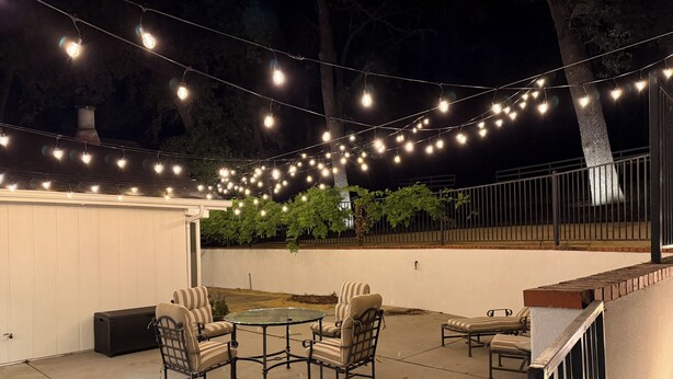 Eine Terrasse im Freien mit Lichterketten, einem runden Glastisch und gepolsterten Stühlen.