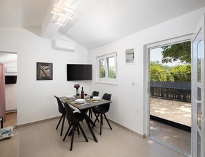 Ein Essbereich mit einem Tisch für vier Personen, einem an der Wand befestigten Flachbildfernseher und einer Glasschiebetür, die auf eine Terrasse im Freien führt.