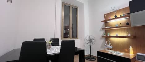 Un espacio habitable pequeño con zona de comedor, estantería de madera empotrada y ventilador de pie.