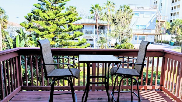 A wooden deck with two chairs and a table, surrounded by lush greenery and buildings in the background. - Be A Nomad | Cozy Beach Retreat | Walk to Shore | Jacksonville Beach (Jacksonville Beach)