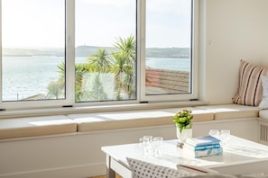 Ein Zimmer mit großem Fenster und Blick auf ein Gewässer, einer Sitzecke mit Kissen, einem Tisch mit Büchern und Gläsern sowie einer Vase mit Blumen.