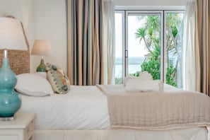 Ein Hotelzimmer mit Bett, Lampe, einem Zierkissen sowie Blick auf Palmen und Wasser durch ein Fenster.