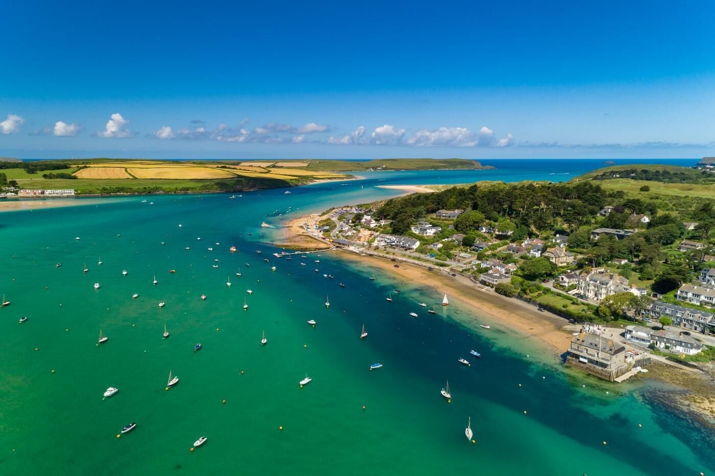 An aerial view of a coastal town with a marina filled with boats, surrounded by green fields and a clear blue sky.