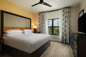 Habitación de hotel con una cama grande, ventilador de techo, televisión y ventana con cortinas estampadas.