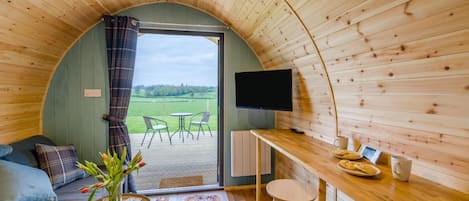 A cozy interior with a curved wooden ceiling, a seating area with a sofa and chairs, a small table with a vase of flowers, a TV mounted on the wall, and a view of a green field through a glass door.
