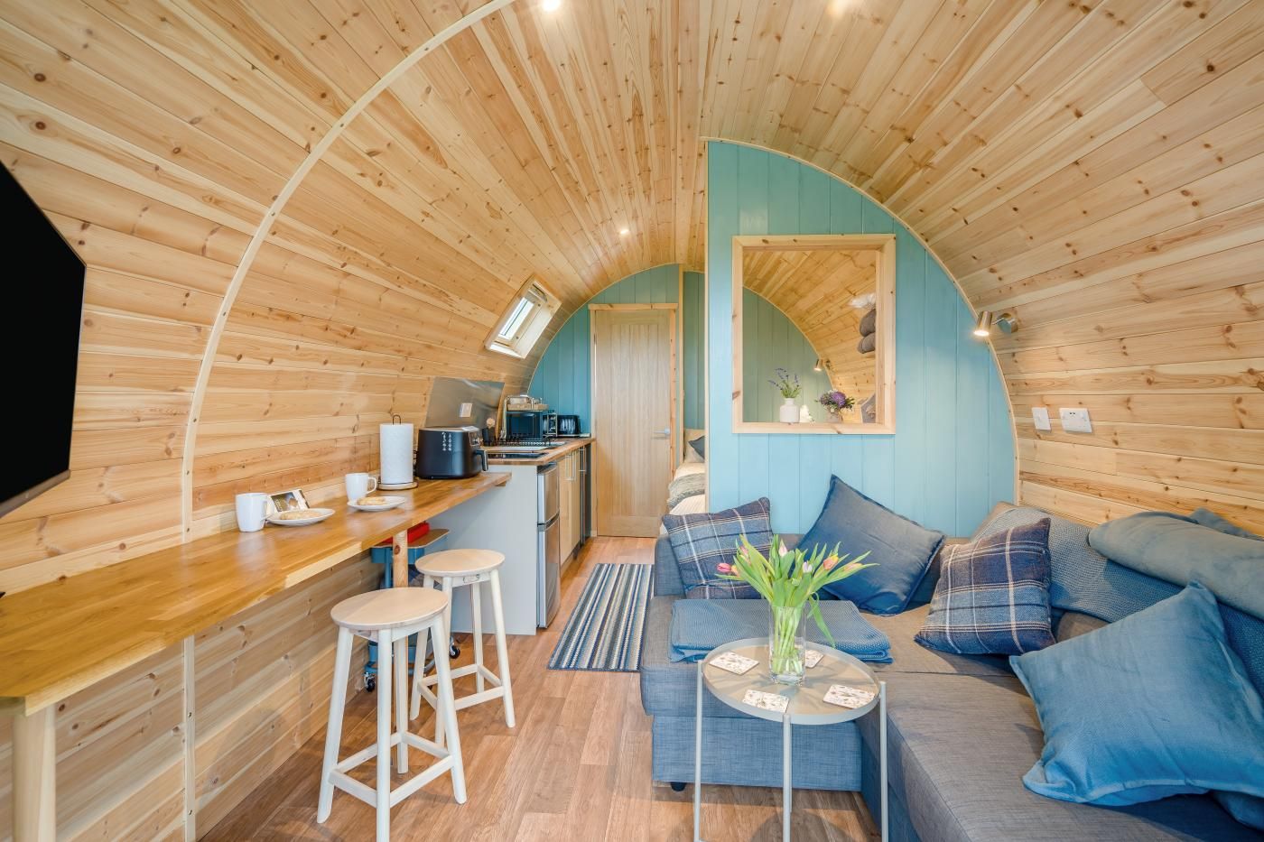 A cozy interior with a wooden ceiling, a kitchenette, a seating area with a sofa and cushions, and a small table with a vase.