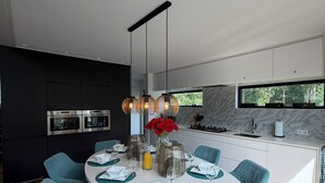 Une cuisine moderne dotée d’appareils électroménagers intégrés, un coin repas avec des chaises de couleur turquoise et une table ronde, et une grande fenêtre donnant sur un espace verdoyant.