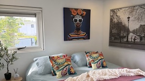 Une chambre à coucher avec un lit, des oreillers, une couverture, une fenêtre donnant sur un beau paysage et des œuvres d’art accrochées aux murs.