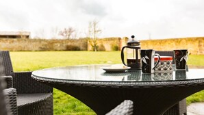 Un tavolo rotondo con una caffettiera, due tazze decorate e un piatto, apparecchiato all'aperto con sedie.