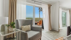 A modern living room with a grey armchair, a glass coffee table, and a view of the outdoors. - Be A Nomad | 2BR Beach Apartment | 1-Min Walk to Sand | Sleeps 4 (Jacksonville Beach)
