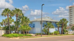 A two-story building with a flat roof, a balcony on the second floor, and a parking lot with palm trees. - Be A Nomad | 2BR Beach Apartment | 1-Min Walk to Sand | Sleeps 4 (Jacksonville Beach)