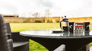 Een ronde tafel met een glazen blad, omringd door rieten stoelen, een koffiepot en bijpassende kopjes.