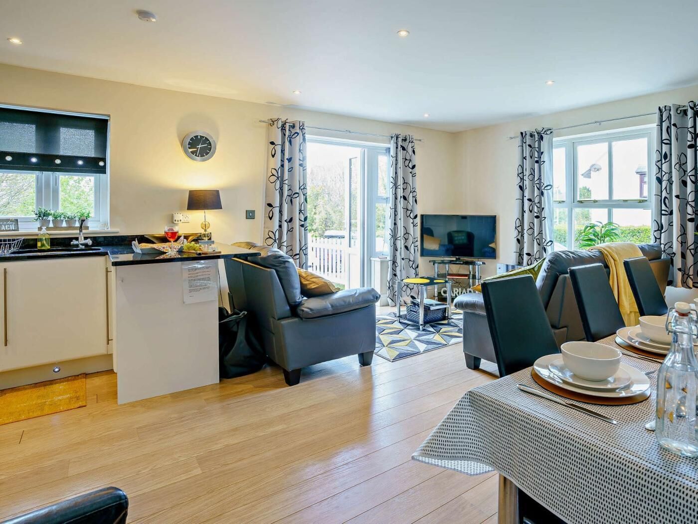 A modern living room with a kitchen area, a dining table set for two, and a television.