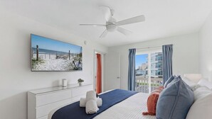 A hotel room with a bed, a TV mounted on the wall, a ceiling fan, a window with curtains, and a view of buildings outside. - Be A Nomad | 3 Bed 3 Bath | 1 Min to Jacksonville Beach | Sleeps 6 (Jacksonville Beach)