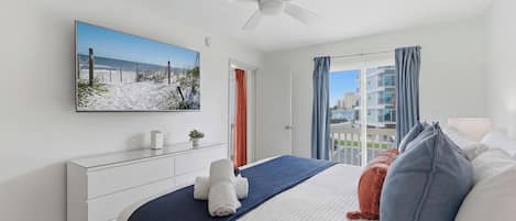 Ett hotellrum med en säng, en tv som är monterad på väggen, en takfläkt, ett fönster med gardiner och utsikt över byggnaderna utanför.