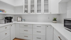 A modern kitchen with white cabinetry, a coffee machine, a framed picture, and a potted plant. - Be A Nomad | Oceanview | Golf Simulator- Sleeps 10 (Jacksonville Beach)