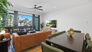 A modern living room with a sofa, dining table, and chairs, large windows with a view, and a ceiling fan. - Be A Nomad | Oceanview | Golf Simulator- Sleeps 10 (Jacksonville Beach)