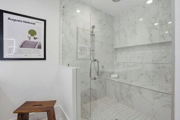 A modern bathroom with a glass-enclosed shower, marble walls, and a framed advertisement on the wall. - Be A Nomad | Oceanview | Golf Simulator- Sleeps 10 (Jacksonville Beach)