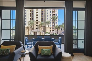 A balcony with two armchairs, a small table, and a view of a hotel building and palm trees. - Be A Nomad | Oceanview | Golf Simulator- Sleeps 10 (Jacksonville Beach)