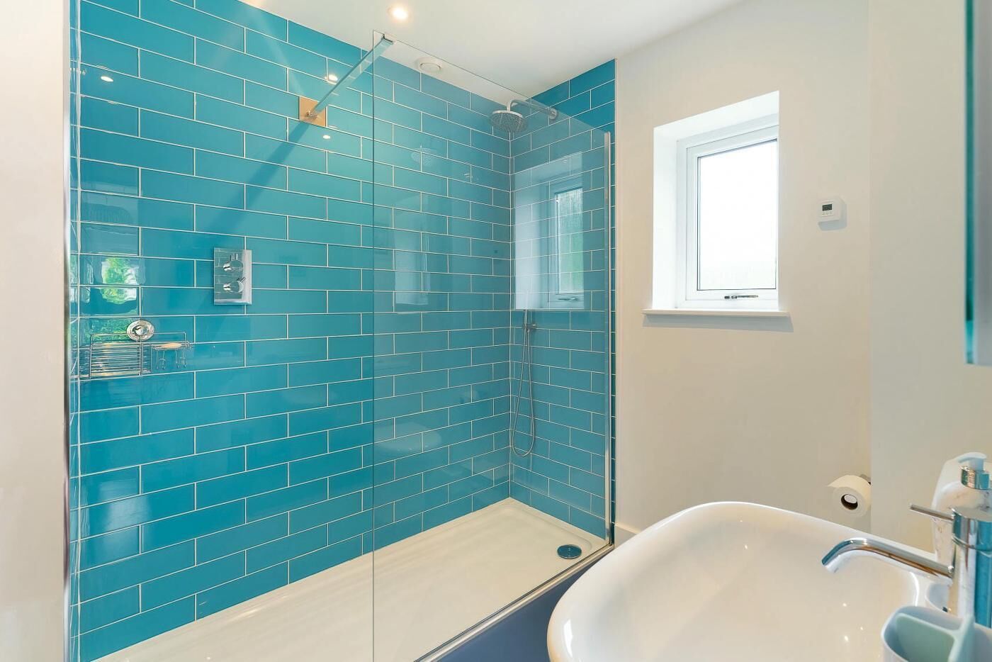 A bathroom with blue tiles, a glass shower enclosure, a white sink, and a window.