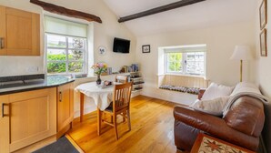 A kitchen with wooden cabinets, a dining table with chairs, a built-in bench with cushions, a television mounted on the wall, and a window with a view of greenery. - 1 Bed in Troutbeck (oc-lr027) (Troutbeck)