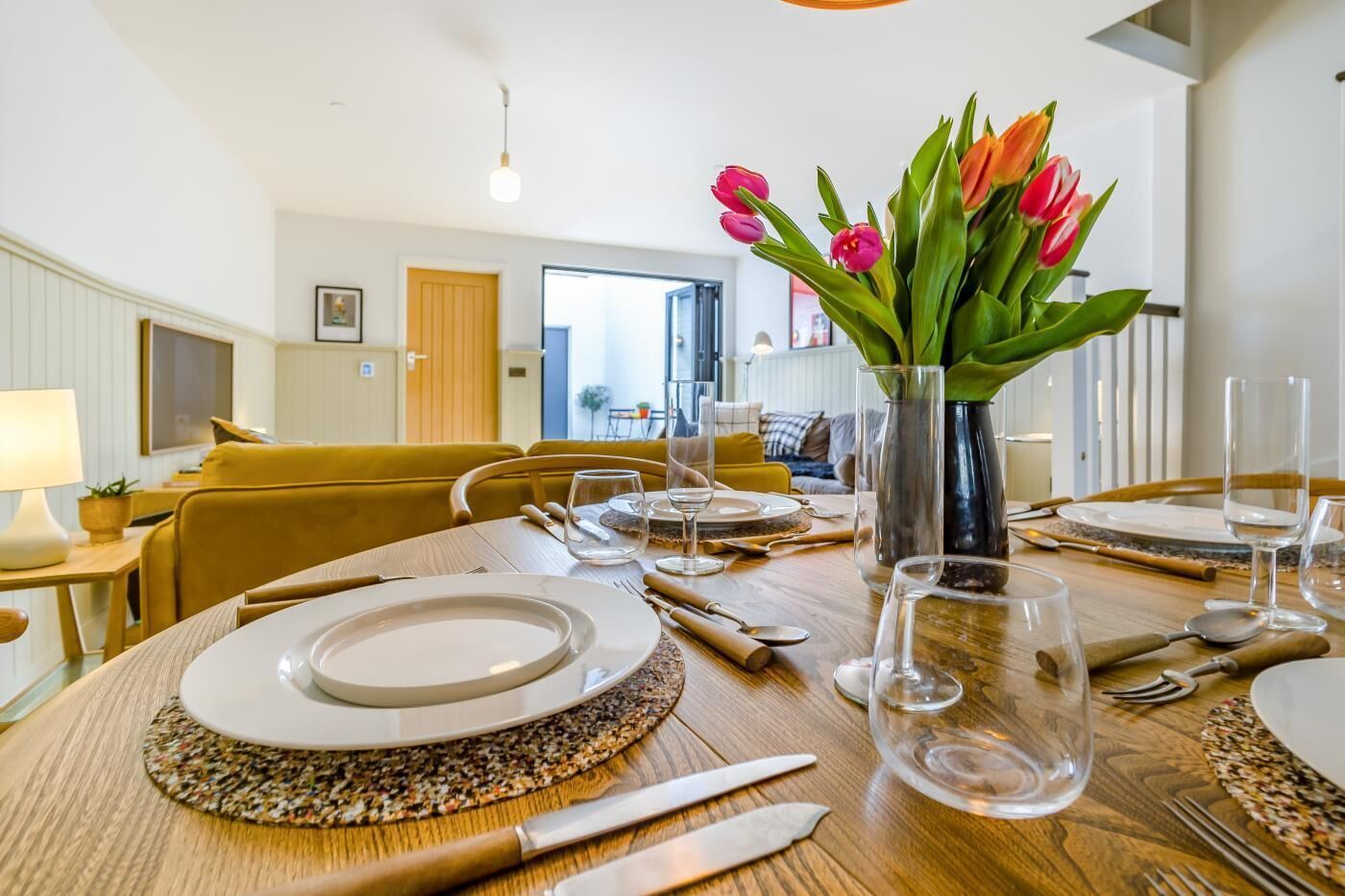 A dining area with a round table set for four, a vase with tulips, and a view into the adjacent living room.