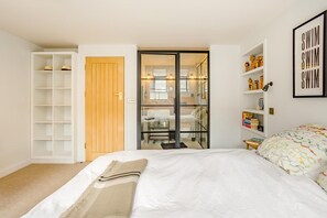 Een slaapkamer met een bed, een kledingkast, een badkamer zichtbaar door een glazen deur, en een plank met boeken en decoratieve objecten.