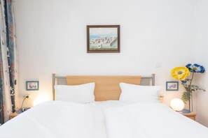 Hotelkamer met houten hoofdeinde, witte beddengoed, een ingelijst beeld op de muur, nachtkastje met lamp en vaas met bloemen.
