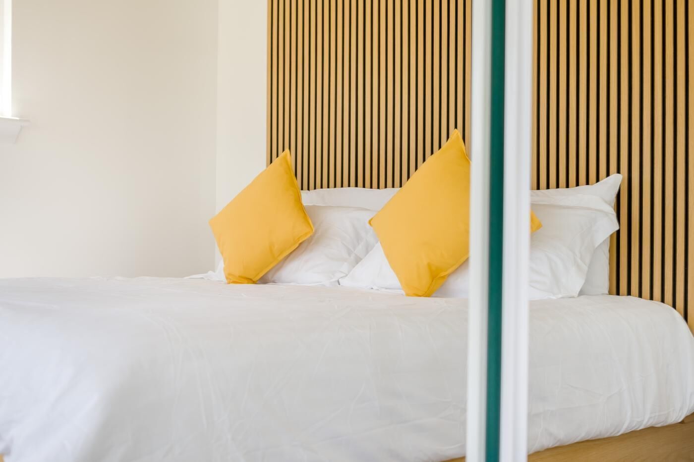 A bed with white bedding and two yellow pillows, a wooden slat headboard, and a green vertical element.