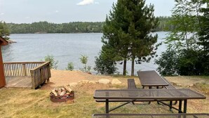 A lakeside outdoor area with a picnic table and lounge chairs, a wooden deck, and a fire pit. - 2bedroom home, lake access, snowmobile trail access, ac/fire place,  Indian Lake (Indian lake)