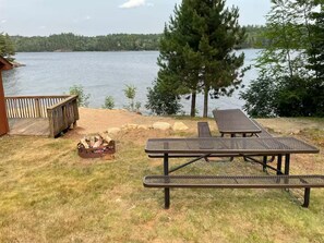 A lakeside outdoor area with a picnic table and lounge chairs, a wooden deck, and a fire pit. - 2bedroom home, lake access, snowmobile trail access, ac/fire place,  Indian Lake (Indian lake)