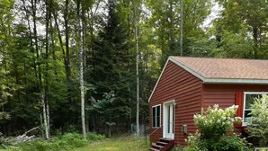 A red wooden cabin with a small porch, surrounded by a grassy yard and dense forest. - 2bedroom home, lake access, snowmobile trail access, ac/fire place,  Indian Lake (Indian lake)