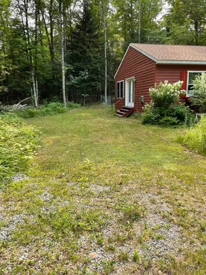 A red wooden cabin with a small porch, surrounded by a grassy yard and dense forest. - 2bedroom home, lake access, snowmobile trail access, ac/fire place,  Indian Lake (Indian lake)