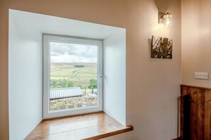 Ein Zimmer mit Fensterblick auf eine ländliche Landschaft, einer hölzernen Bank und einer wandmontierten Lampe.