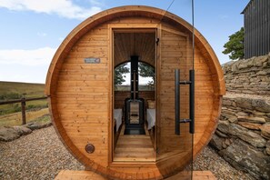 Eine Holzsauna mit Schild, das zum Schuhe ausziehen auffordert, einer Glastür und einer Steinwand im Hintergrund.