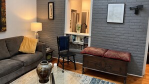 Un salon avec un mur en briques grises, un canapé, un fauteuil, une petite table sur laquelle se trouve un vase, et une bibliothèque.