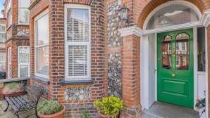 Une maison en briques rouges avec une porte verte, une fenêtre avec des volets et un banc à l’extérieur.