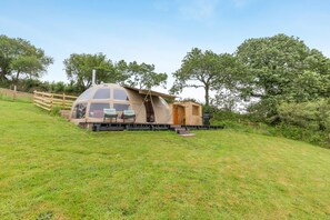 A dome-shaped tent with a wooden exterior, a small attached cabin, and a grassy hillside setting.